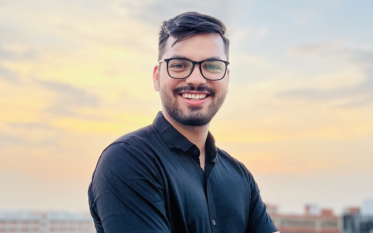 Smiling man in black shirt with glasses against sunset sky background, standing outdoors.