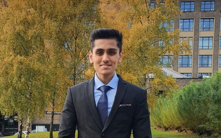 Man in suit standing outdoors with autumn trees and buildings in background.