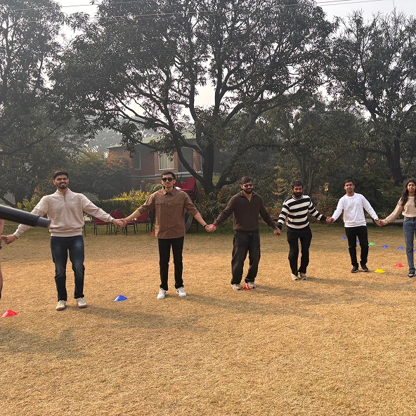 Group of people holding hands outdoors, forming a circle on grass, with trees and a house in the background during daylight.